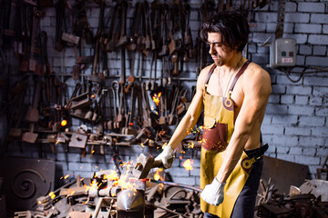 Authentic blacksmith man forges a metal product in dark indoors studio