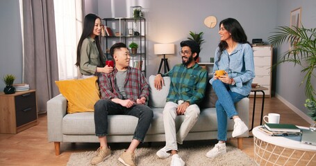 Joyful young mixed-races family couples sitting on sofa in modern living room and laughing in good mood. Asian and Hindu men and women at home drinking coffee or tea and speaking, friends concept