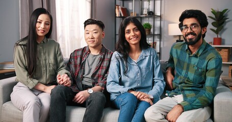  Portrait of Asian and Hindu young female and male friends sit together on sofa in apartment looking at camera and smiling. Multi-ethnic couples with smiles on faces, positive emotions, Leisure concept