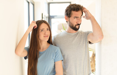 Wall Mural - young couple feeling puzzled and confused, scratching head and looking to the side