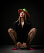 Naughty arrogant young sexy woman in jacket, fishnet tights stockings, pink high-heeled shoes and hat sits squatted looking at upper corner over dark background. Sexual games, bdsm, glamour concept
