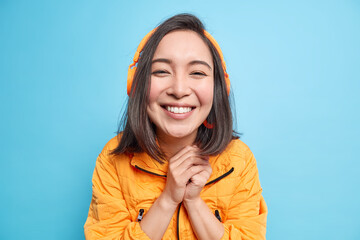 Wall Mural - Portrait of happy beautiful Asian woman keeps hands together smiles broadly enjoys good sound with modern headset listens music from playlist wears orange jacket isolated over blue background