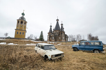 April, 2021 - Nyonoksa. Old Russian rusty cars on the background of wooden Orthodox churches. Russia, Arkhangelsk region 