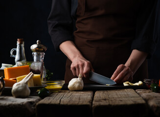 Wall Mural - Chef slices garlic for preparing a dish of meat and salad. On the background of ingredients, cheeses, olive oil and spices. Recipes and food preparation, illustration for the menu book