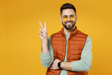 Wall Mural - Young smiling happy friendly cheerful fun caucasian man 20s in orange vest mint sweatshirt glasses show victory v-sign gesture isolated on yellow background studio portrait. People lifestyle concept