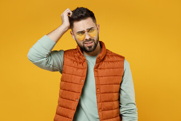 Wall Mural - Young puzzled pensive troubled thoughtful man 20s wearing orange vest mint sweatshirt glasses looking aside sctratch hold head isolated on yellow color background studio portrait. Lifestyle concept.