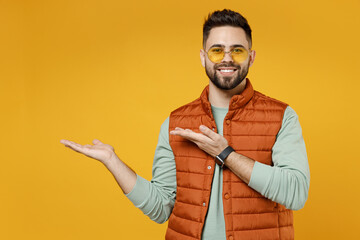 Wall Mural - Young smiling happy fun man 20s in orange vest mint sweatshirt glasses point arms hands aside on copy space area mock up workspace isolated on yellow background studio portrait. Advertisement concept.