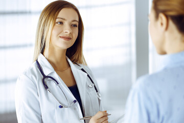 Wall Mural - Young woman-doctor and her patient are discussing patient's current health examination, while standing together in a hospital office. Female physician is writing some marks, using a clipboard. Perfect