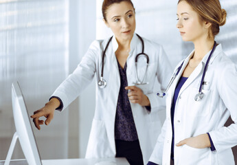 Wall Mural - Two female physicians are discussing medical therapy, while standing at the table in a clinic office. Doctors use pc computer at work. Teamwork in medicine