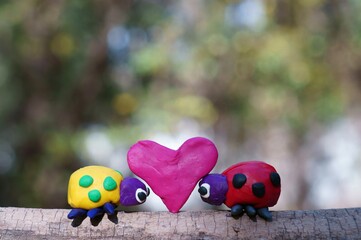 Canvas Print - Two ladybugs made of plasticine in the forest on a colored background. Between them is a small red heart.