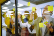© maxbelchenko - Group of young modern people working on project together. Business colleagues writing on sticky note on glass wall with coworkers standing by in office. Startup Business. Planning, analysis.