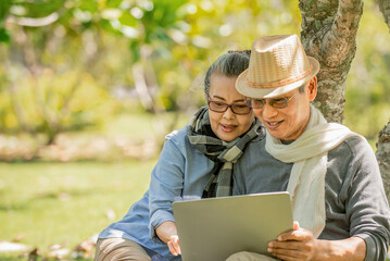 Wall Mural - Senior asian couple  sitting under the tree  at park  using  laptop computer while holding coffee cup