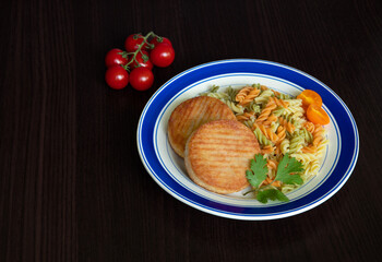 Two cutlets with pasta, greens and cherry tomatoes on plate. Dark background, isolated, copy space.