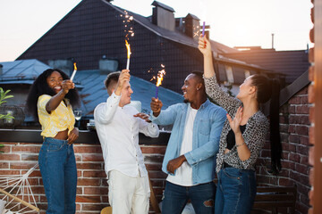 Wall Mural - Joyful multiracial friends smiling, chatting and using sparklers during rooftop party. Young hipster people enjoying leisure time spending together.