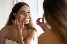 Happy Millennial Teenage Latin Woman Look In Mirror In Bathroom Apply Nourishing Moisturizing Cream For Health Glowing Face Skin. Smiling Young Hispanic Female Do Beauty Facial Procedures In Bath.