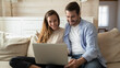 Happy millennial multiracial man and woman relax on sofa at home look at laptop screen shopping online together. Smiling young couple rest on couch in living room use computer browsing internet.