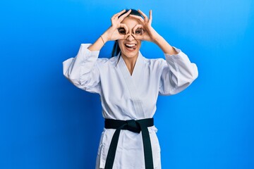 Wall Mural - Beautiful brunette young woman wearing karate fighter uniform with black belt doing ok gesture like binoculars sticking tongue out, eyes looking through fingers. crazy expression.