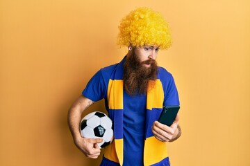 Canvas Print - Redhead man with long beard football hooligan cheering game holding smartphone clueless and confused expression. doubt concept.