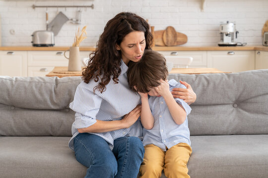 Young mother comfort crying son. Disturbed mom hug small upset preschool boy on couch in living room. Female parent embrace little kid hiding face in hands from abuse or pain. Depressed child concept