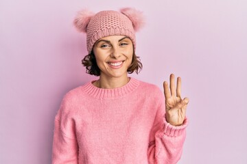 Wall Mural - Young brunette woman wearing cute wool cap showing and pointing up with fingers number three while smiling confident and happy.