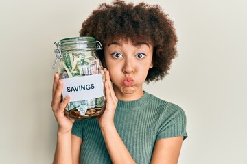 young hispanic girl holding jar with savings puffing cheeks with funny face. mouth inflated with air