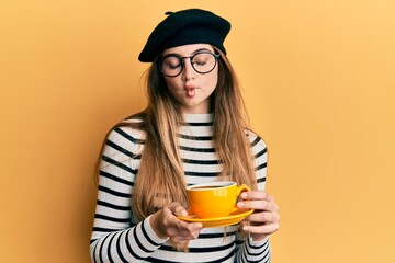 Wall Mural - Young caucasian woman wearing french style drinking a cup of coffee making fish face with mouth and squinting eyes, crazy and comical.