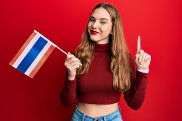 Poster - Young blonde woman holding thailand flag smiling with an idea or question pointing finger with happy face, number one