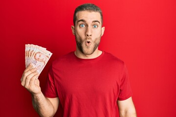 Poster - Young caucasian man holding 10 singapore dollars banknotes scared and amazed with open mouth for surprise, disbelief face