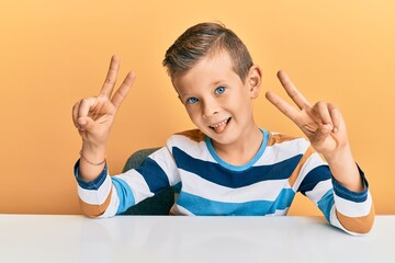 Adorable caucasian kid wearing casual clothes sitting on the table smiling with tongue out showing fingers of both hands doing victory sign. number two.