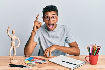 Canvas Print - Young handsome african american man painter sitting palette and art manikin pointing finger up with successful idea. exited and happy. number one.