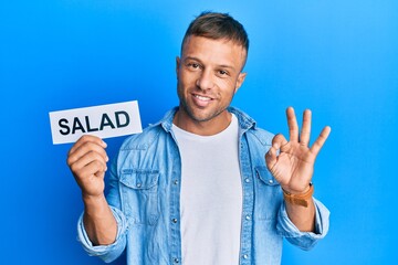 Sticker - Handsome muscle man holding salad word on paper doing ok sign with fingers, smiling friendly gesturing excellent symbol