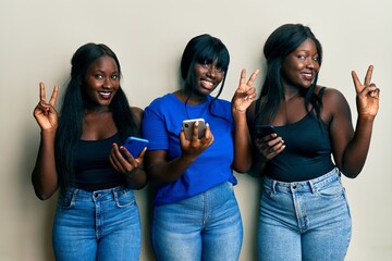 Three young african american friends using smartphone smiling looking to the camera showing fingers doing victory sign. number two.