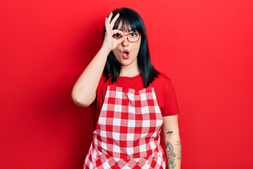 Wall Mural - Young hispanic woman wearing cook apron and glasses doing ok gesture shocked with surprised face, eye looking through fingers. unbelieving expression.