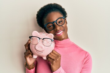 Sticker - Young african american girl holding piggy bank with glasses looking positive and happy standing and smiling with a confident smile showing teeth