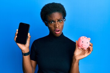 Poster - Young african american girl holding piggy bank and smartphone in shock face, looking skeptical and sarcastic, surprised with open mouth