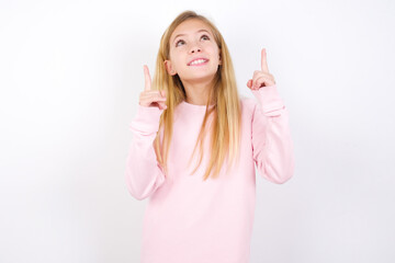 Wall Mural - Successful friendly looking beautiful caucasian little girl wearing pink hoodie over white background exclaiming excitedly, pointing both index fingers up, indicating something.