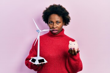 Poster - Young african american woman holding windmill and piggy bank in shock face, looking skeptical and sarcastic, surprised with open mouth