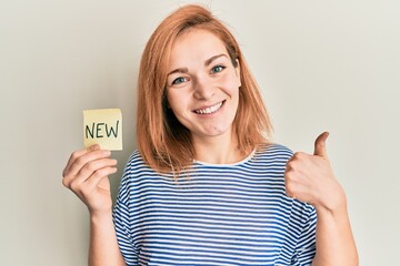 Young caucasian woman holding reminder with new word smiling happy and positive, thumb up doing excellent and approval sign