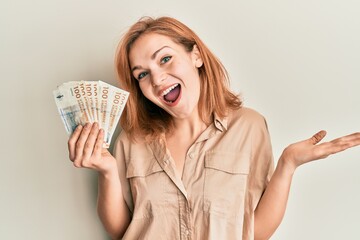 Wall Mural - Young caucasian woman holding 100 danish krone banknotes celebrating achievement with happy smile and winner expression with raised hand