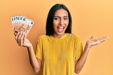 Canvas Print - Young brunette woman holding 50 euro banknotes celebrating achievement with happy smile and winner expression with raised hand