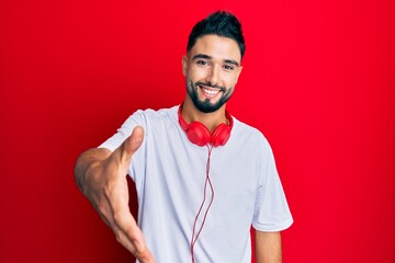 Wall Mural - Young man with beard listening to music using headphones smiling friendly offering handshake as greeting and welcoming. successful business.