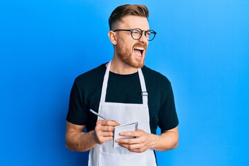 Wall Mural - Young redhead man wearing waiter apron taking order angry and mad screaming frustrated and furious, shouting with anger. rage and aggressive concept.