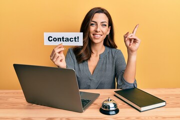 Wall Mural - Young brunette woman working at hotel reception holding contact paper smiling happy pointing with hand and finger to the side