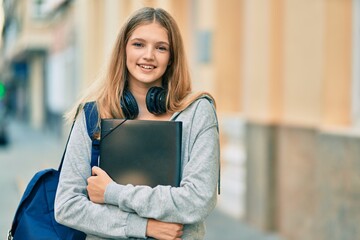 Beautiful caucasian student teenager using headphones holding binder at the city.