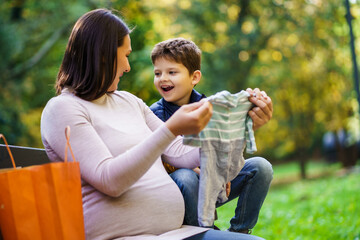 Happy boy and his pregnant mother are are looking at clothes for baby.