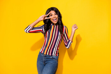 Poster - Portrait of adorable friendly dark skin lady beaming smile fingers show v-sign near eye isolated on yellow color background