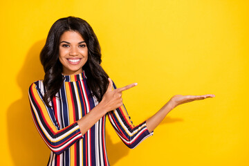 Poster - Photo of charming positive dark skin lady direct finger arm palm hold empty space isolated on yellow color background