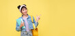 © NaMong Productions - Happy Asian girl with backpack holding books and pointing finger on blank space beside over yellow background. back to school concept.