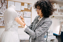 Work Process Of A Tailor In Her Studio. Fashion Designer Sewing Bridal Dress, Pinning Lace On Mannequin.