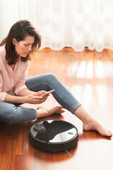 Wall Mural - Young caucasian woman using automatic vacuum cleaner to clean the floor, controlling smart machine housework robot with smart phone at home